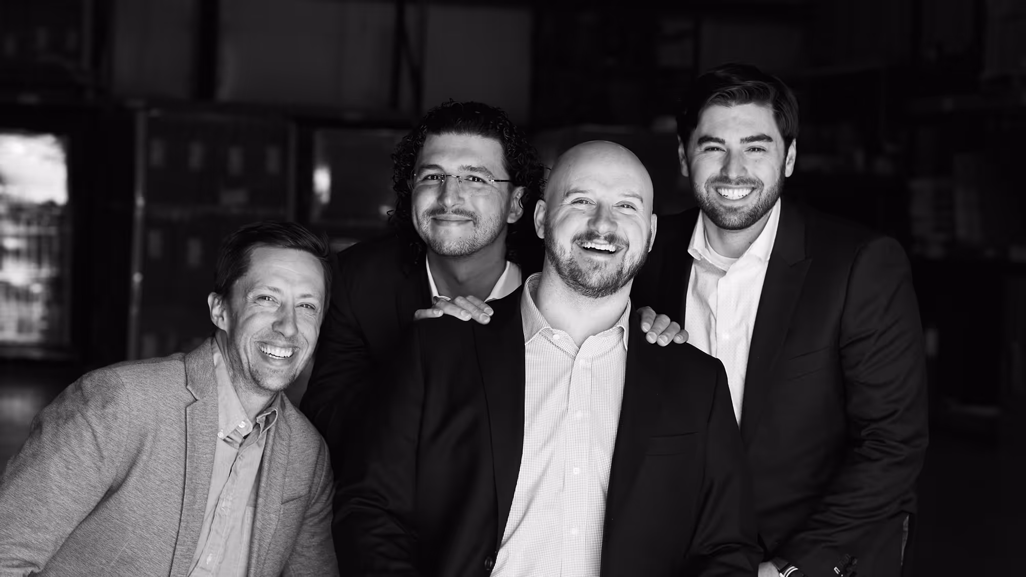Four men in business attire smiling together in a close group photo with a dark background.