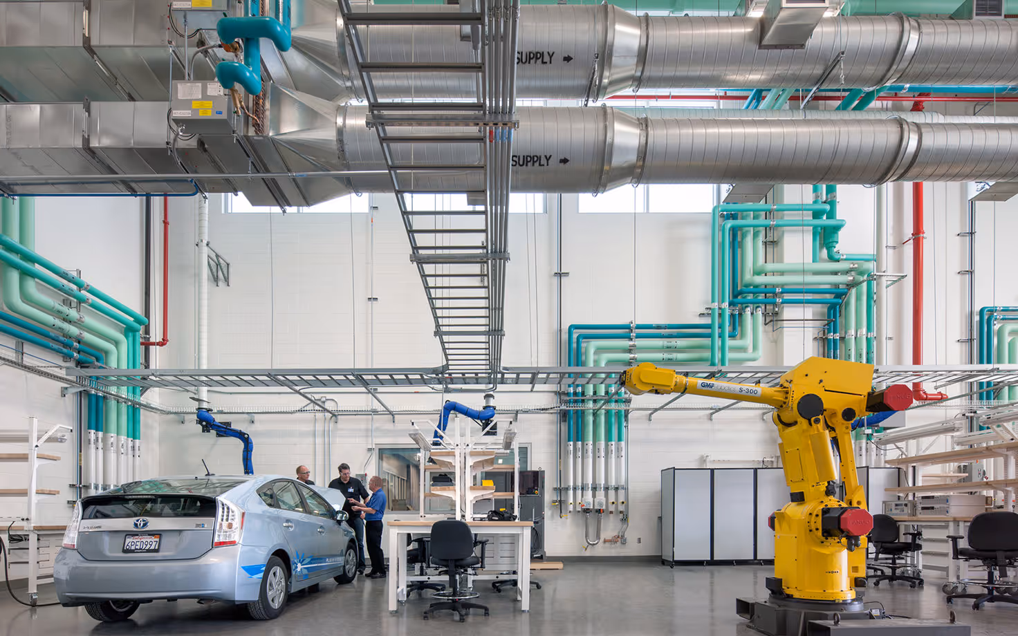 Industrial workshop with a silver Toyota Prius, three men discussing near the car, large yellow robotic arm, and colorful pipes on walls and ceiling.