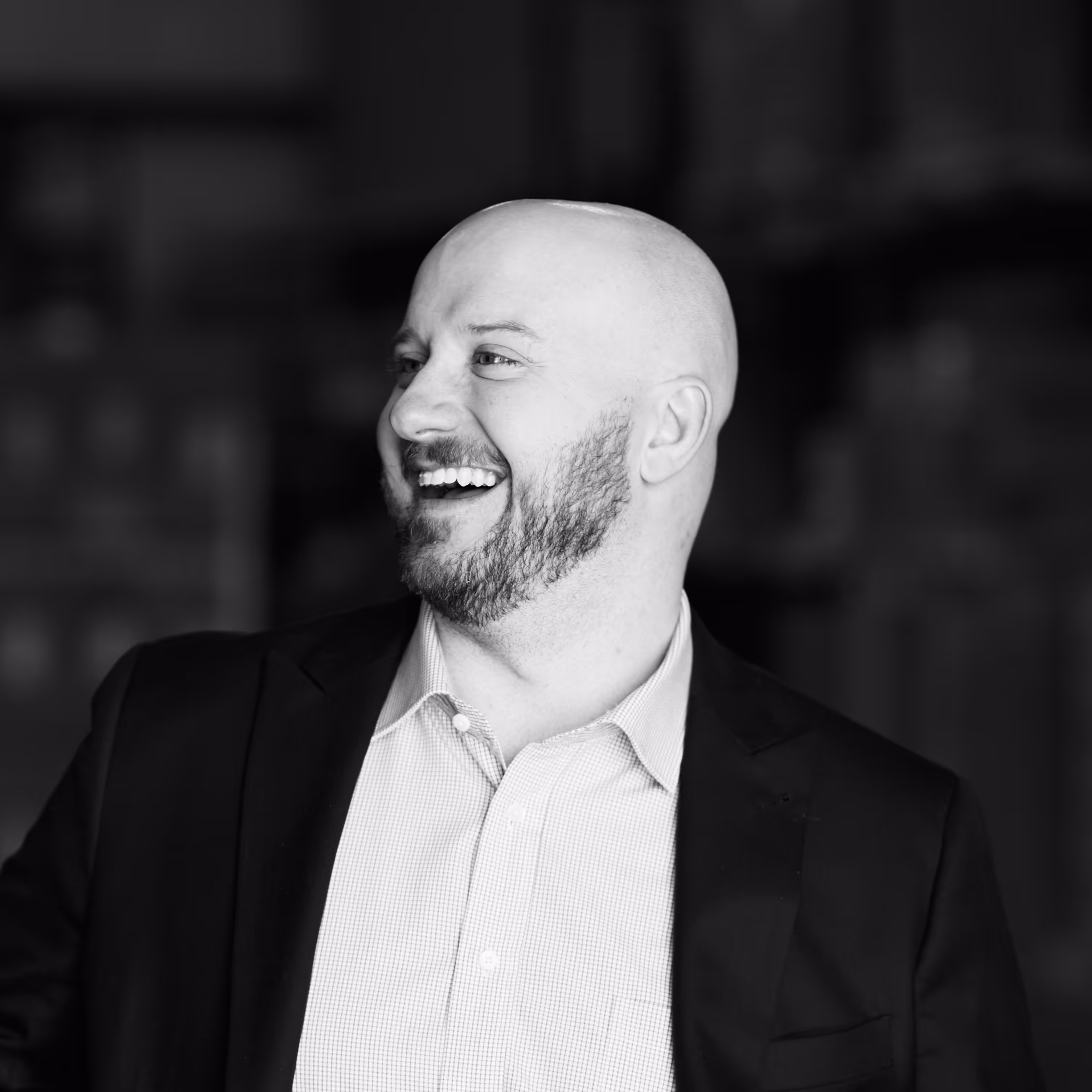 Smiling bald man with a beard wearing a suit jacket and checkered shirt, looking to the side.