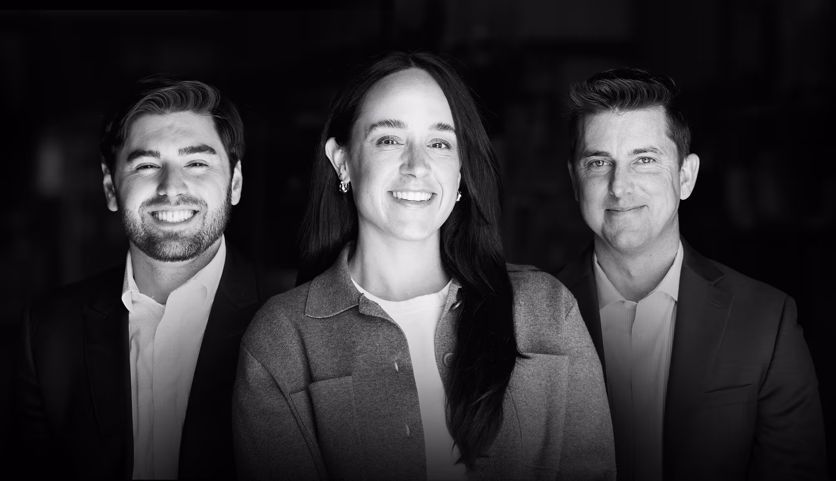 Three professional adults smiling, two men in suits on either side of a woman wearing a buttoned shirt and earrings.