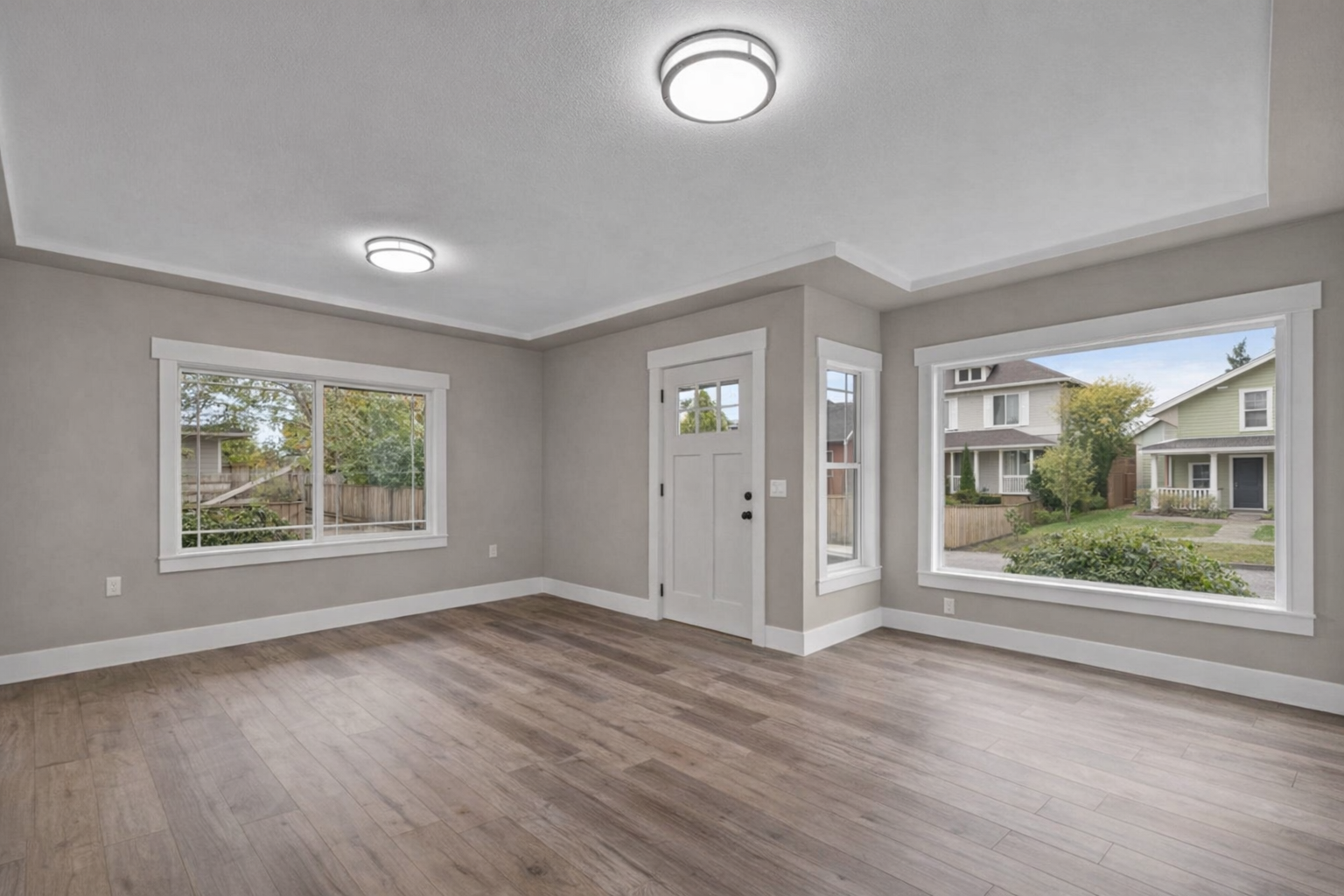 Empty room with wood flooring, beige walls, white trim, a white front door, and large windows showing a view of houses and greenery outside.