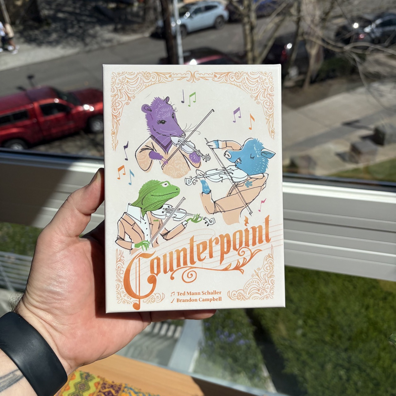 A hand holding the game "Countepoint." The game is in an off-white box, with color animals playing stringed instruments and the game logo.