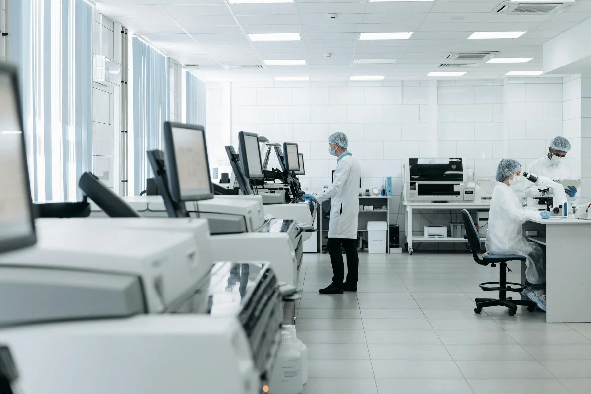 Scientists work in a bright, modern lab. One stands near machines, while two use microscopes at a table. The setting is clean and orderly.