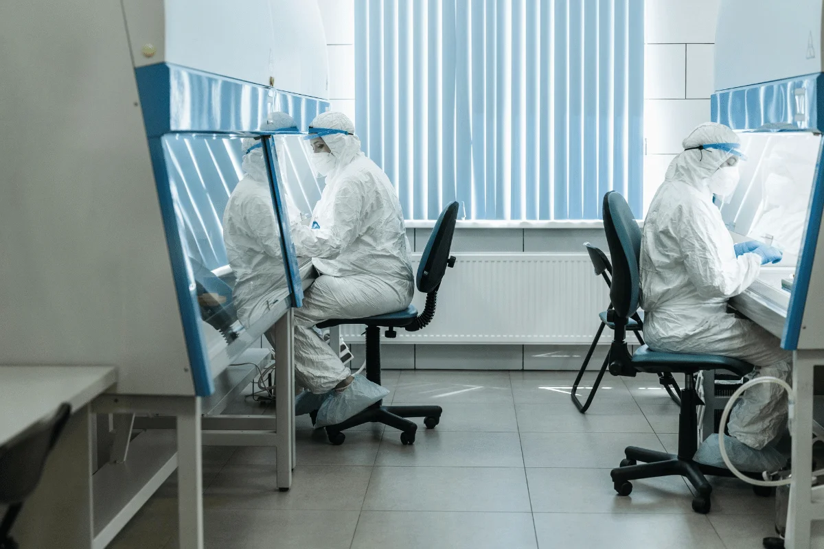 Two scientists in protective suits work in a lab, seated at separate stations with blue accents, under bright lighting and blinds in the background.