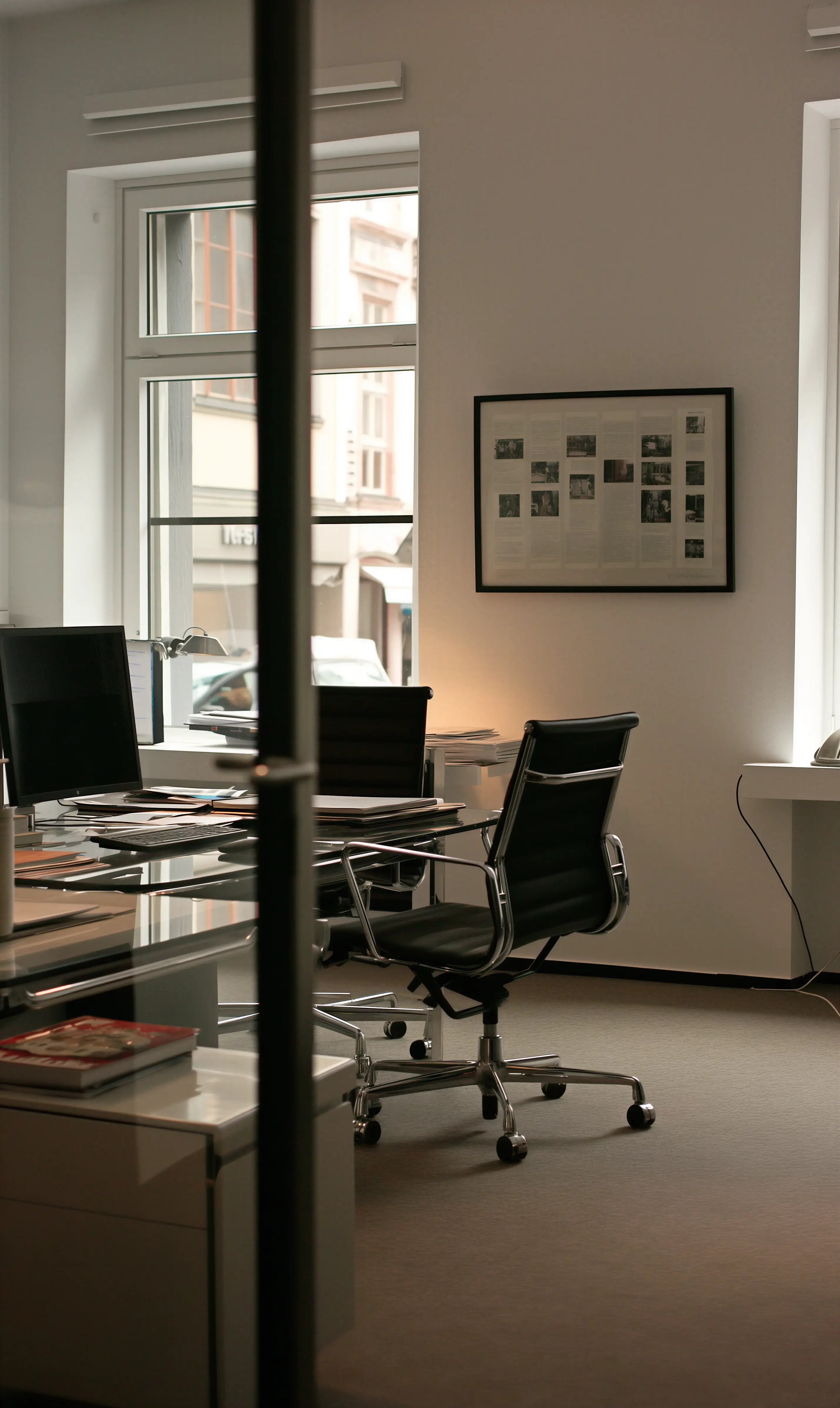 Modern office interior with glass desk, black office chairs, a computer monitor, and a framed document on the wall.