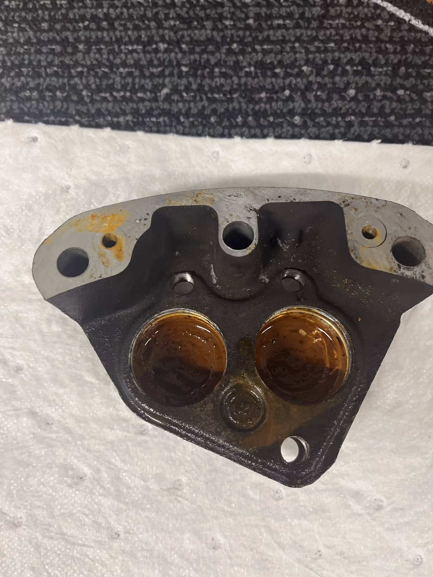 Close-up of a metal engine cylinder head with two valve openings showing oil residue, placed on a white surface.