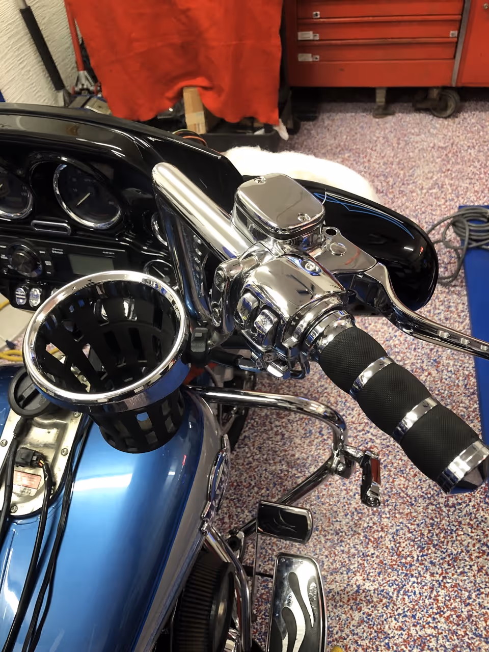 Close-up of a blue motorcycle's right handlebar featuring chrome controls, a black foam grip with silver bands, and a cup holder attached near the fuel tank.