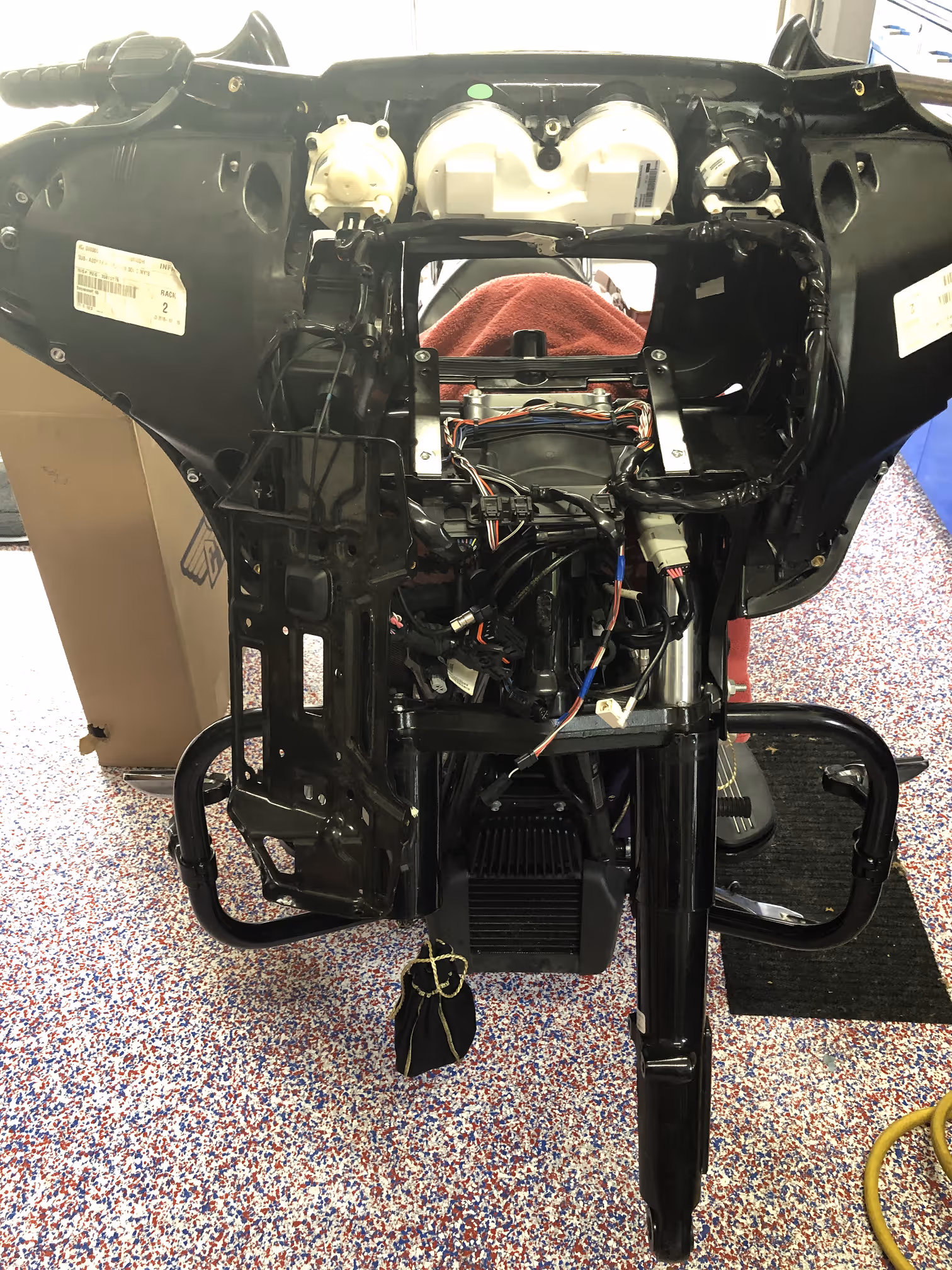 Front view of a partially disassembled motorcycle dashboard and wiring assembly on a speckled red, white, and blue floor.