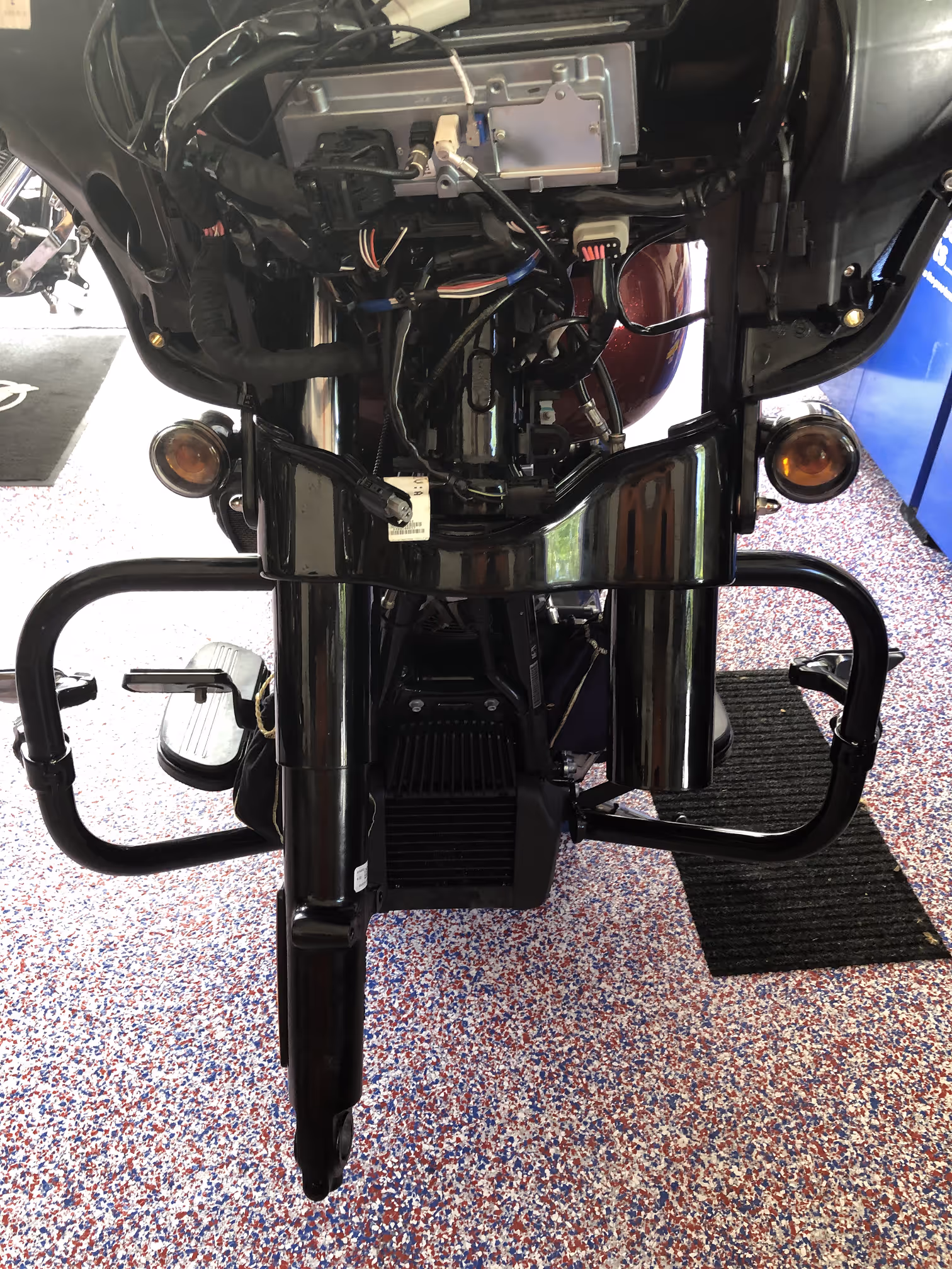Front view of a motorcycle's black frame and exposed wiring on a multicolored speckled floor.