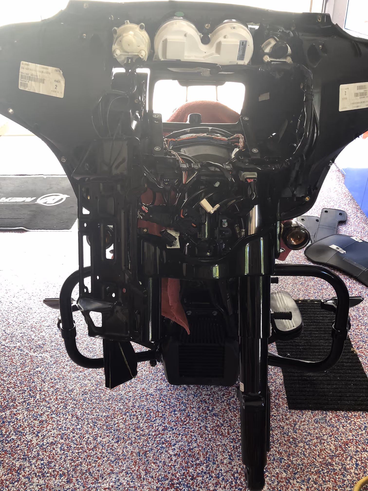 Rear view of a partially disassembled motorcycle frame with exposed wiring and components, resting on a speckled floor.