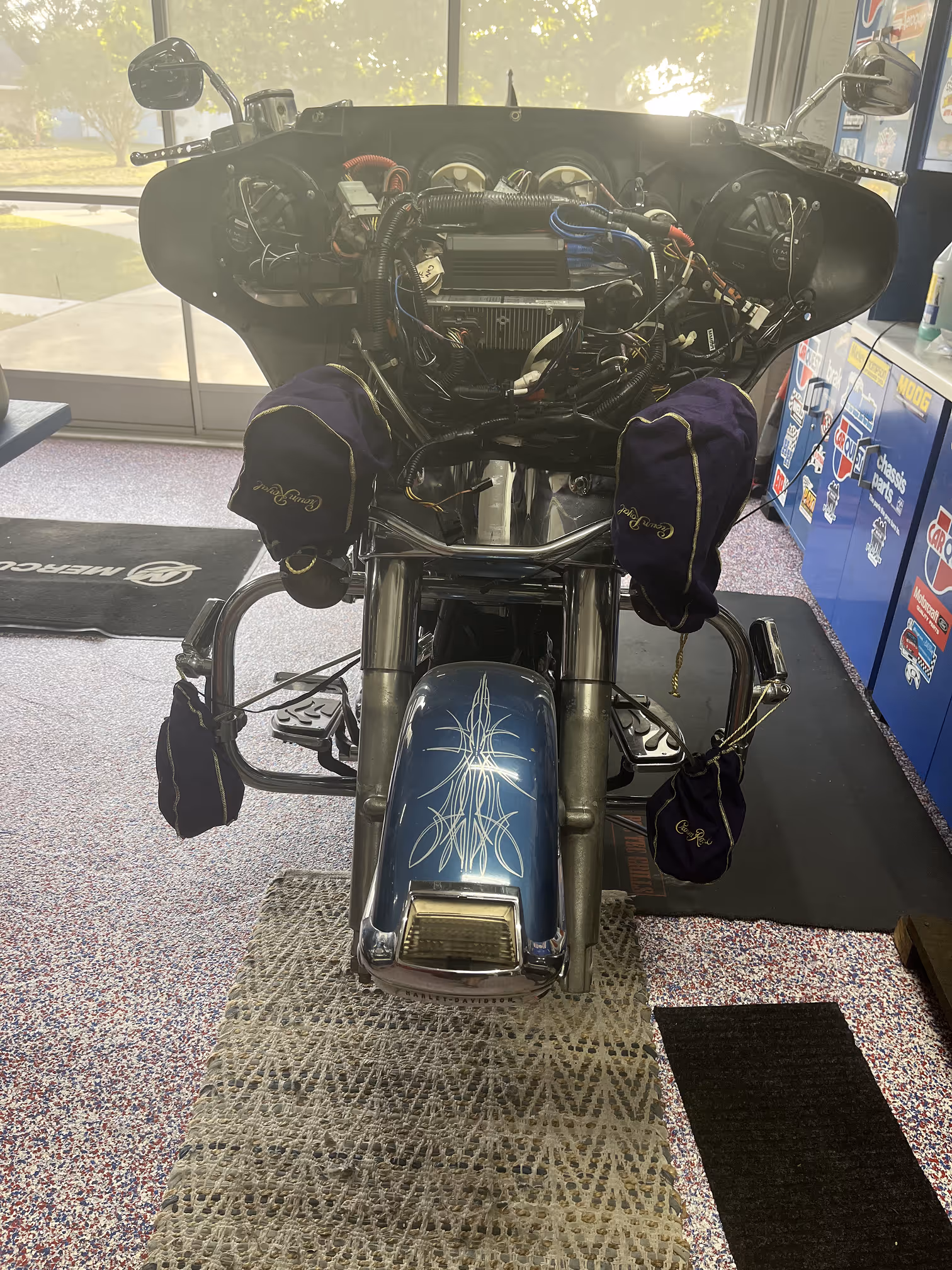 Front view of a Harley-Davidson motorcycle with exposed wiring and blue fender featuring white pinstripe detailing, indoors on patterned floor mats.