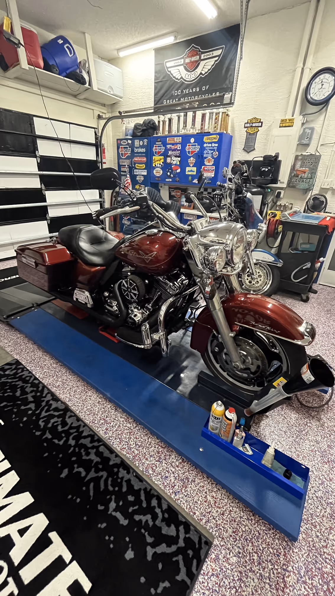 Maroon Harley-Davidson motorcycle with chrome details inside a garage with Harley-Davidson banners and blue tool cabinet covered in stickers.