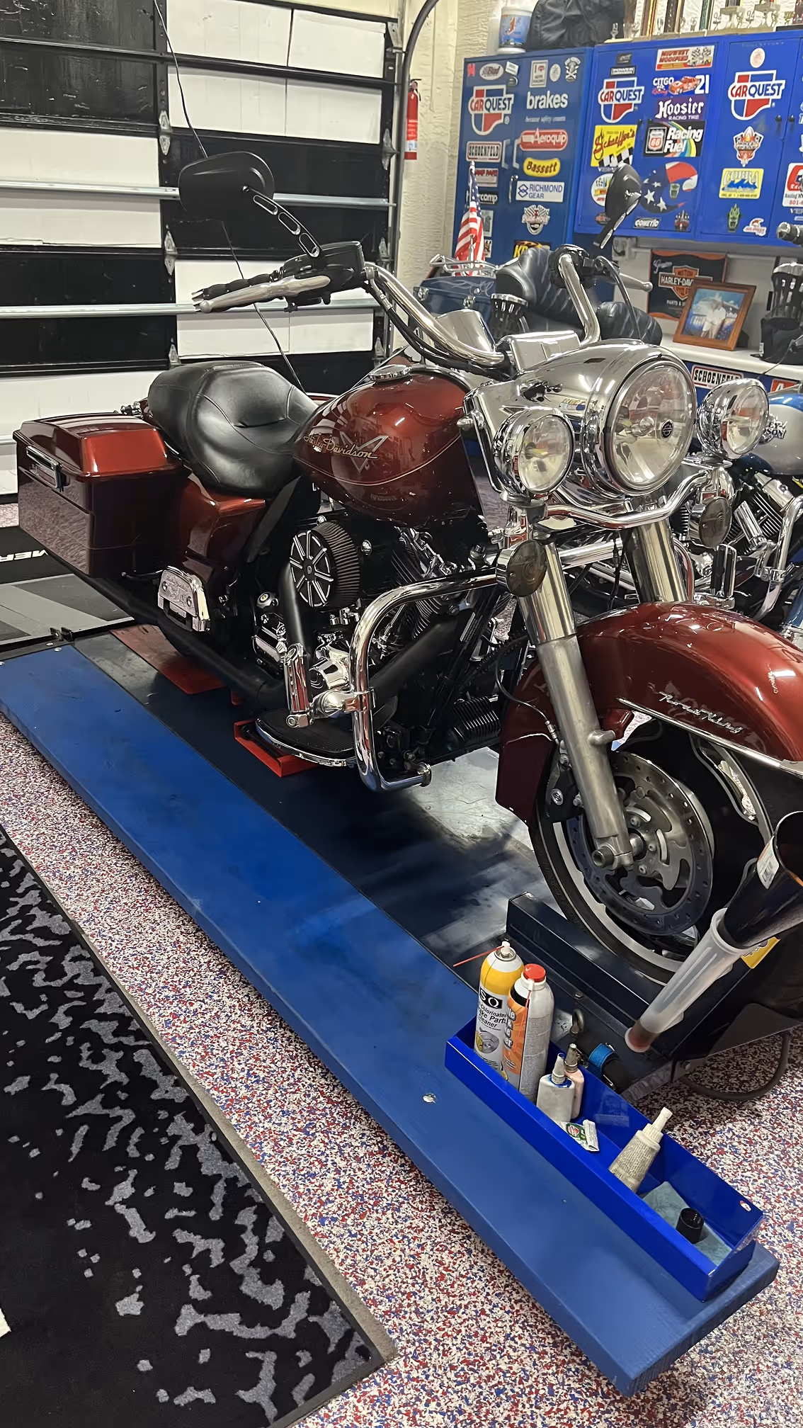 Maroon Harley-Davidson motorcycle parked on a blue lift inside a garage with tool cabinets and stickers in the background.