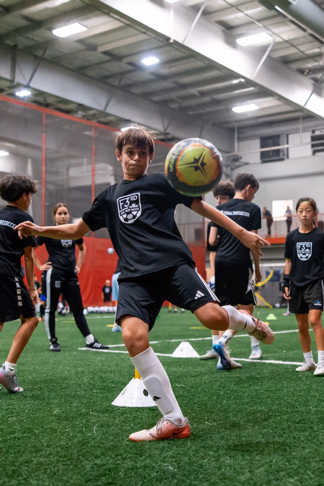 Young soccer player in black uniform controlling a colorful ball indoors while others watch on artificial turf.