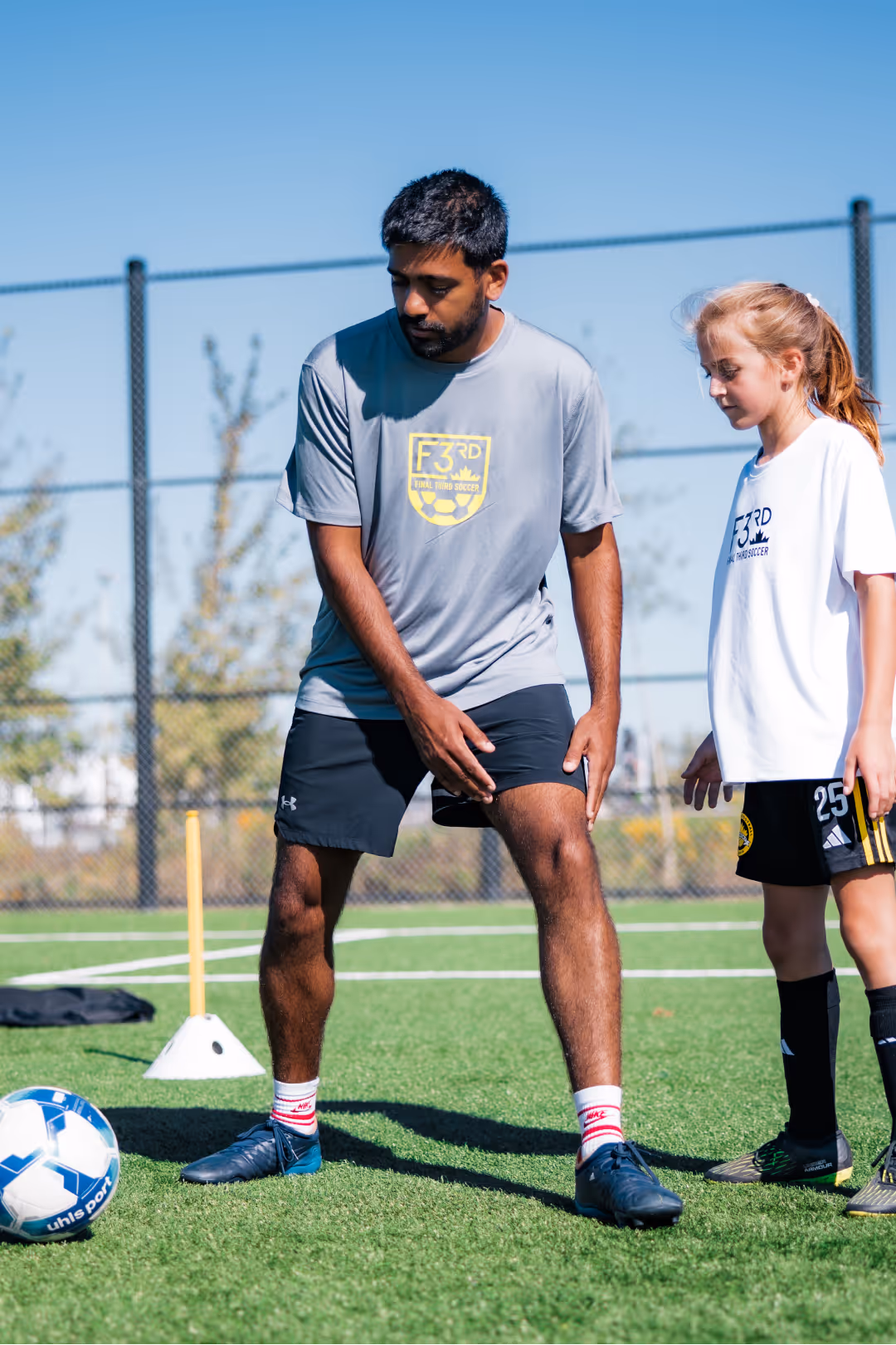 Man in gray soccer shirt demonstrating a soccer move to a young girl in white soccer shirt on a green field.