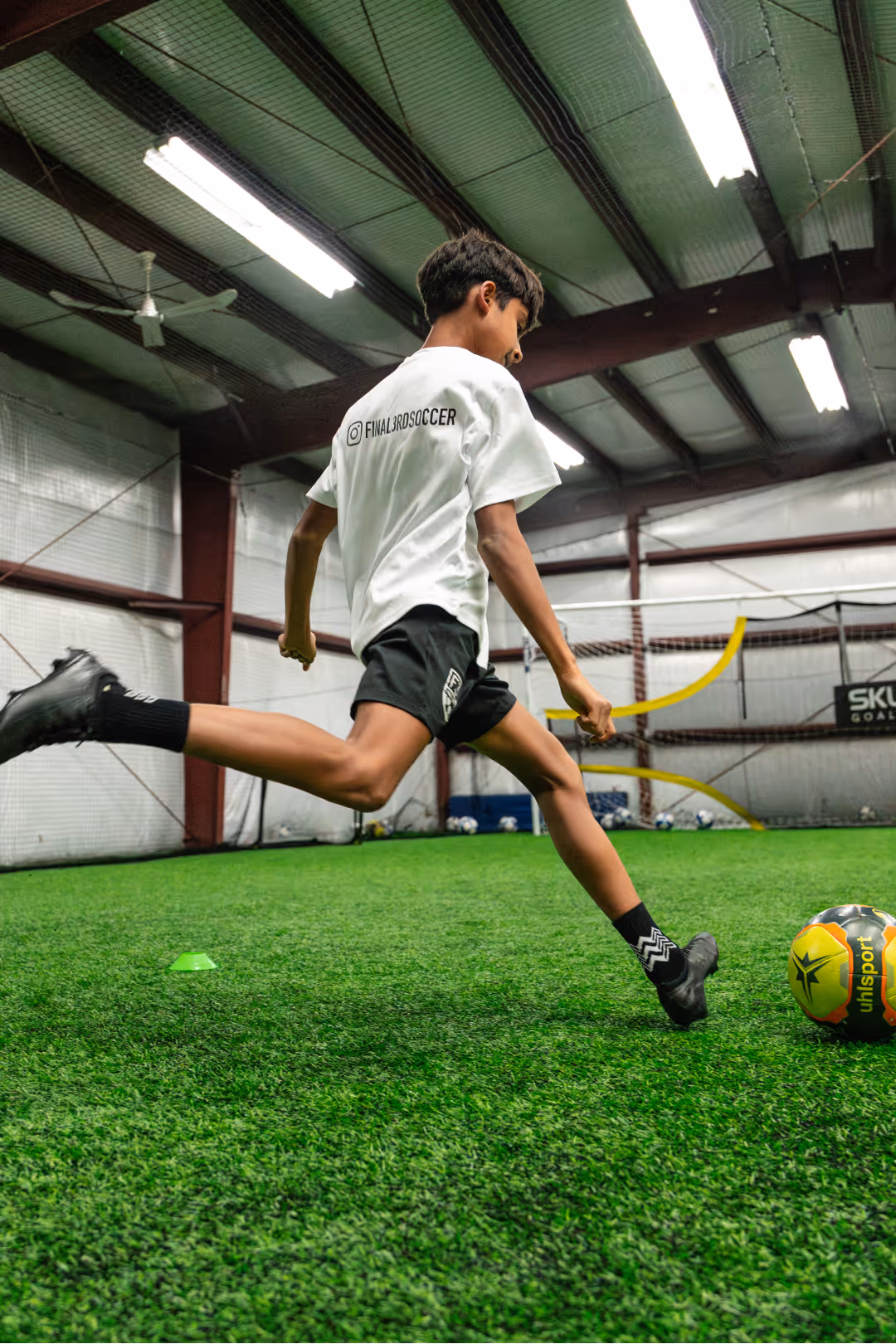 Young boy in a white t-shirt and black shorts kicking a soccer ball indoors on artificial turf.