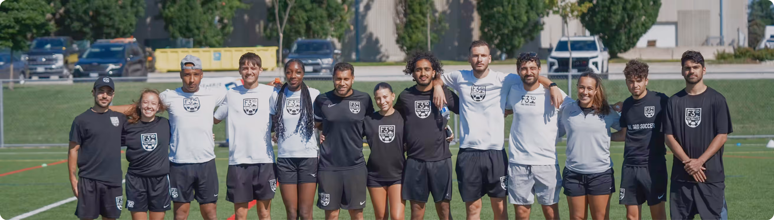 Group of twelve young soccer coaches standing arm in arm on a grass field wearing black and white sportswear with F3 3rd Soccer logos.