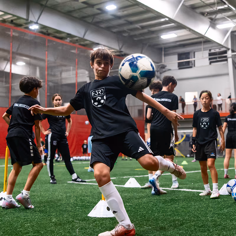 Children practicing soccer skills indoors on artificial turf while wearing black F3RD All Pro Soccer shirts.
