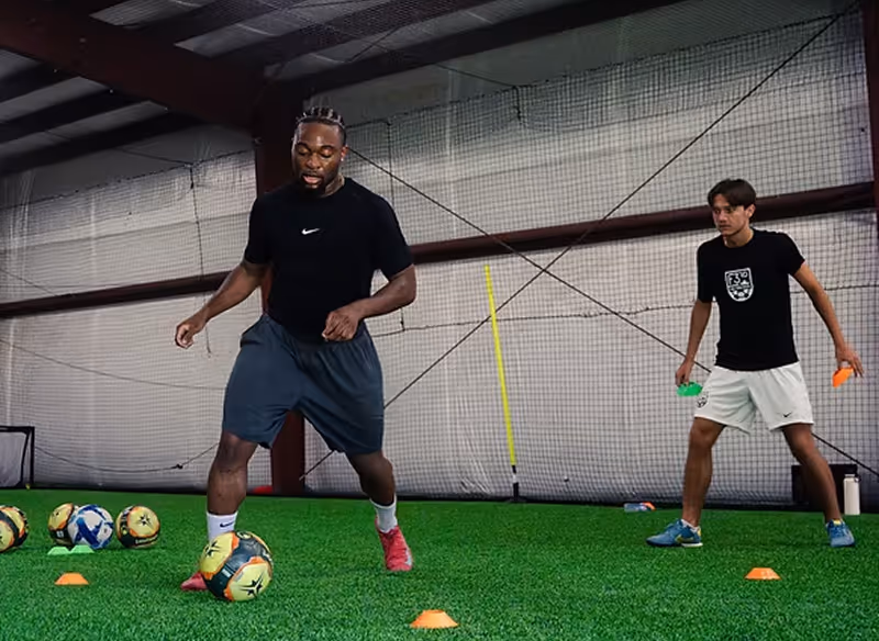 Two men training indoors on artificial turf with several soccer balls and orange cones; one man is dribbling a ball while the other watches holding small training cones.