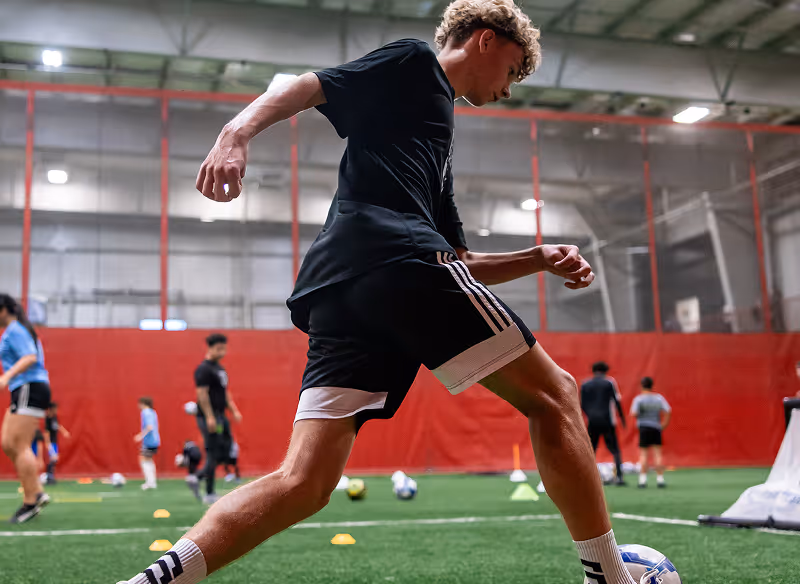 Young male soccer player in black sportswear kicking a soccer ball on an indoor turf field with other players practicing in the background.