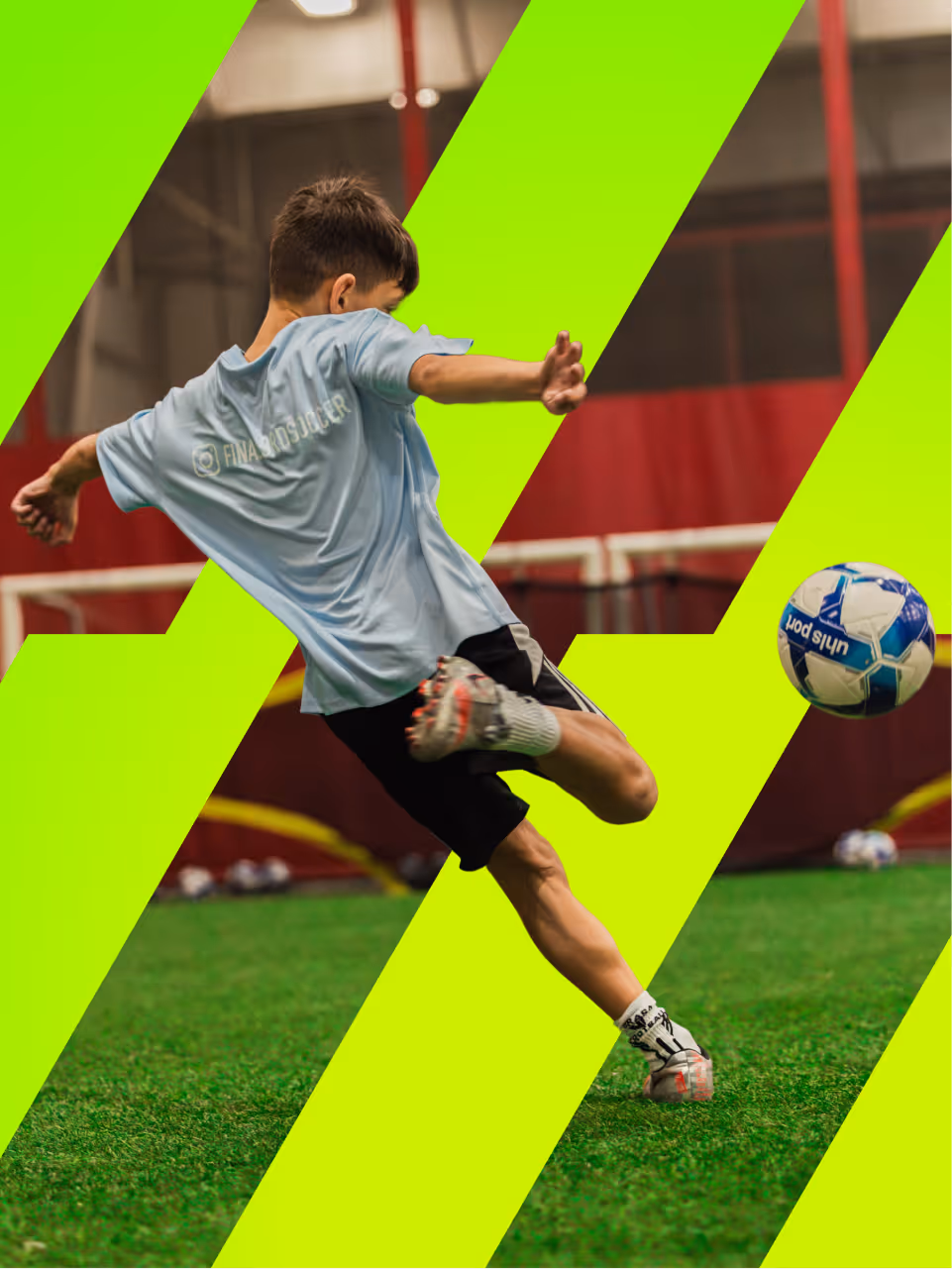 Young boy in blue shirt and black shorts kicking a soccer ball indoors on green turf.