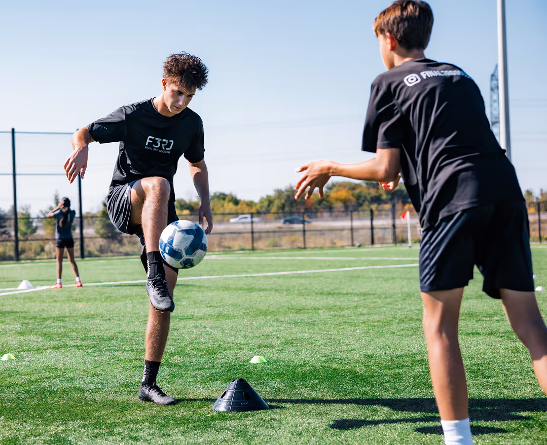 Two teenage boys in black sportswear practicing soccer on a green field, one juggling a soccer ball with his knee.