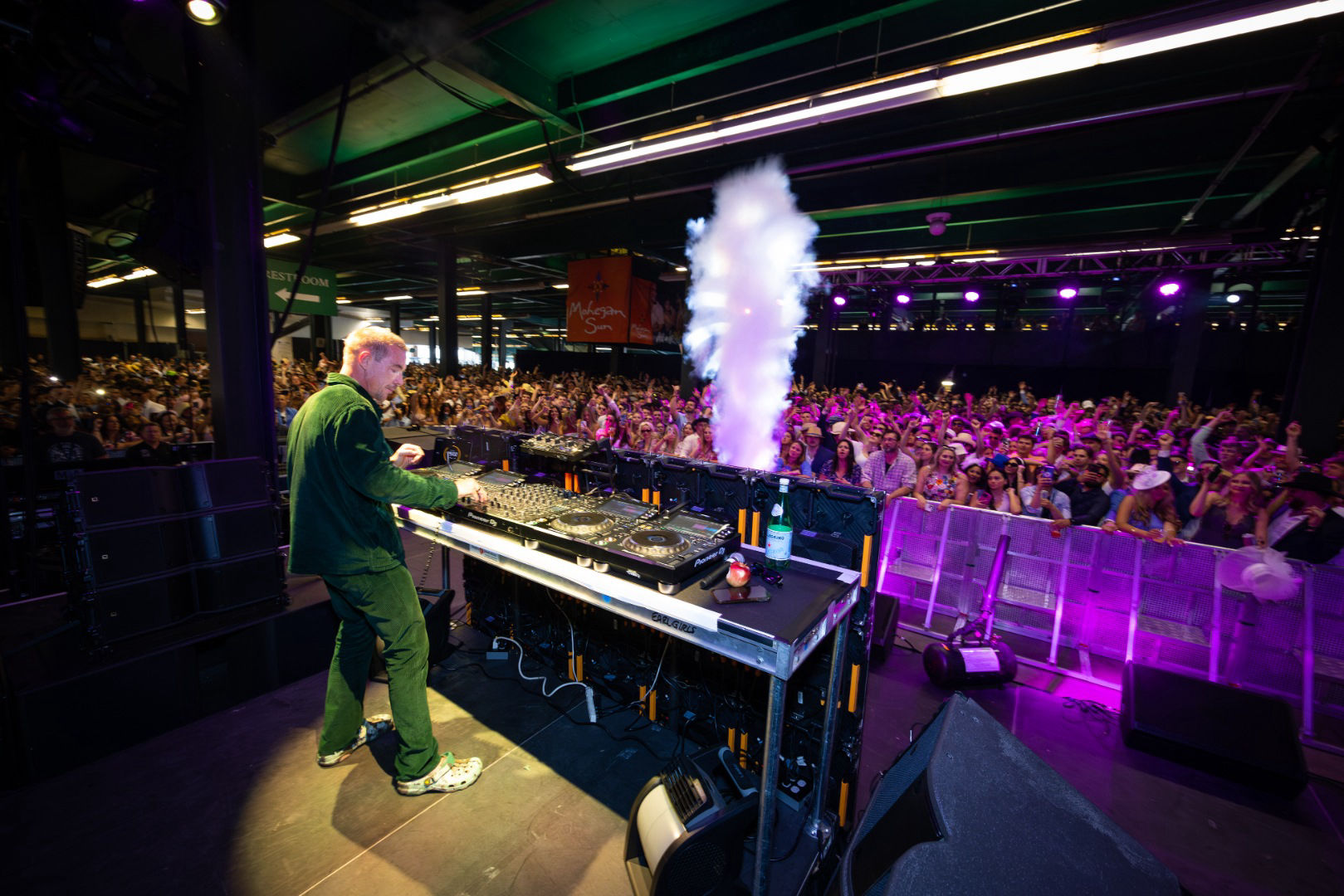 DJ performing on stage with crowd and smoke effects at an indoor music event.