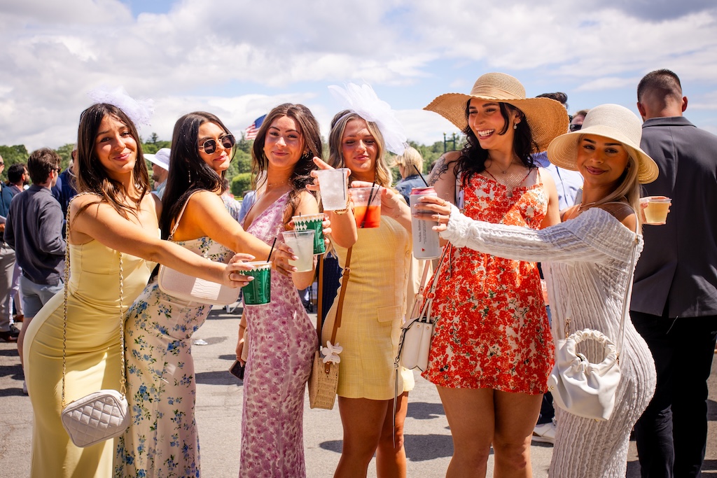 Six women in summer dresses and hats holding drinks outdoors in a festive gathering.