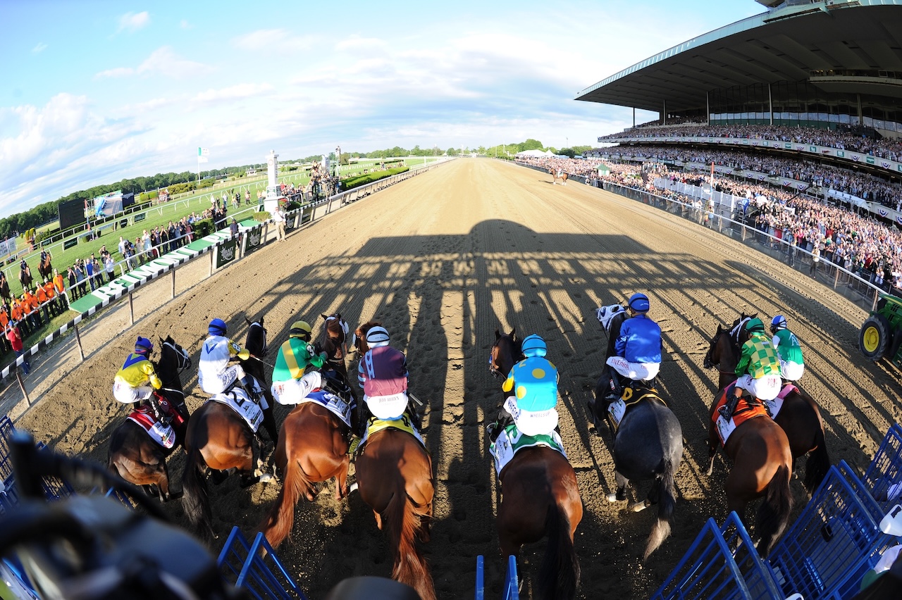 Jockeys on horses lined up in starting gates at a racetrack with spectators in stands.