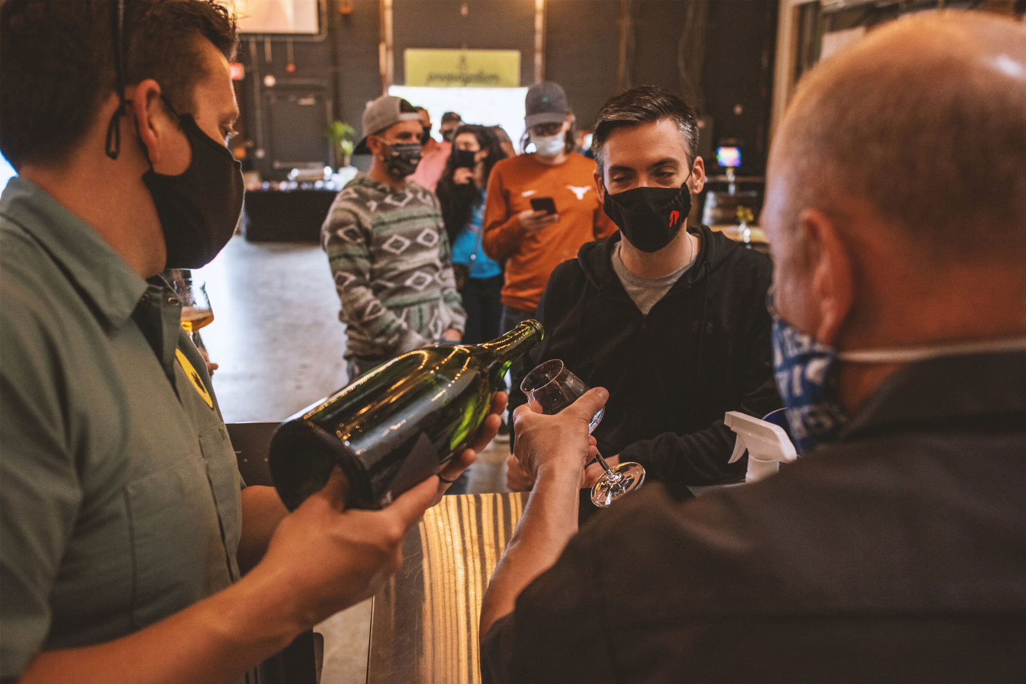 A masked server pours wine into a glass for a customer at a bar. People in the background wear masks, suggesting a safe, social atmosphere.