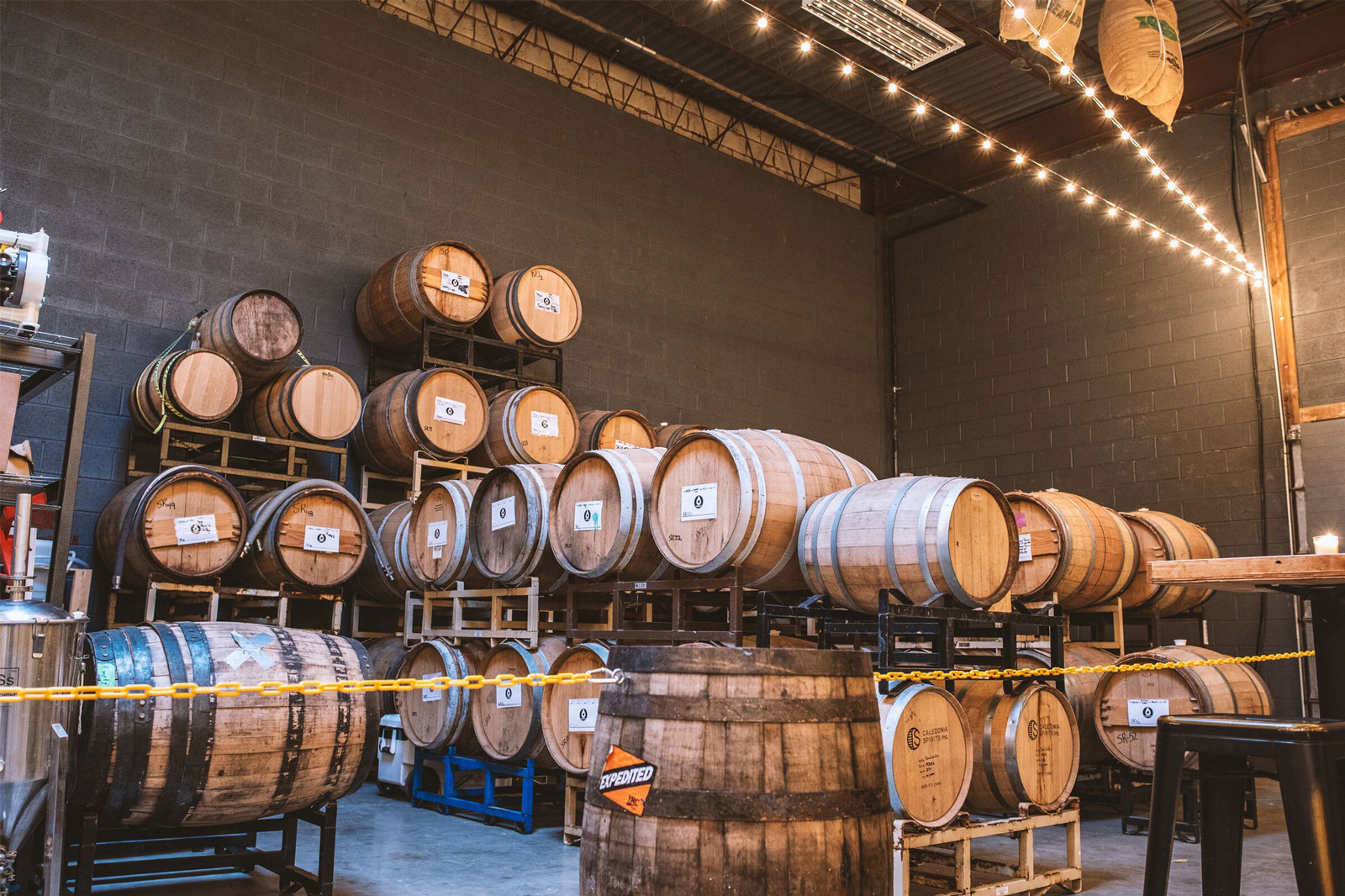 A dimly lit warehouse with wooden barrels stacked against dark gray walls. Warm string lights add a cozy ambience to the industrial setting.