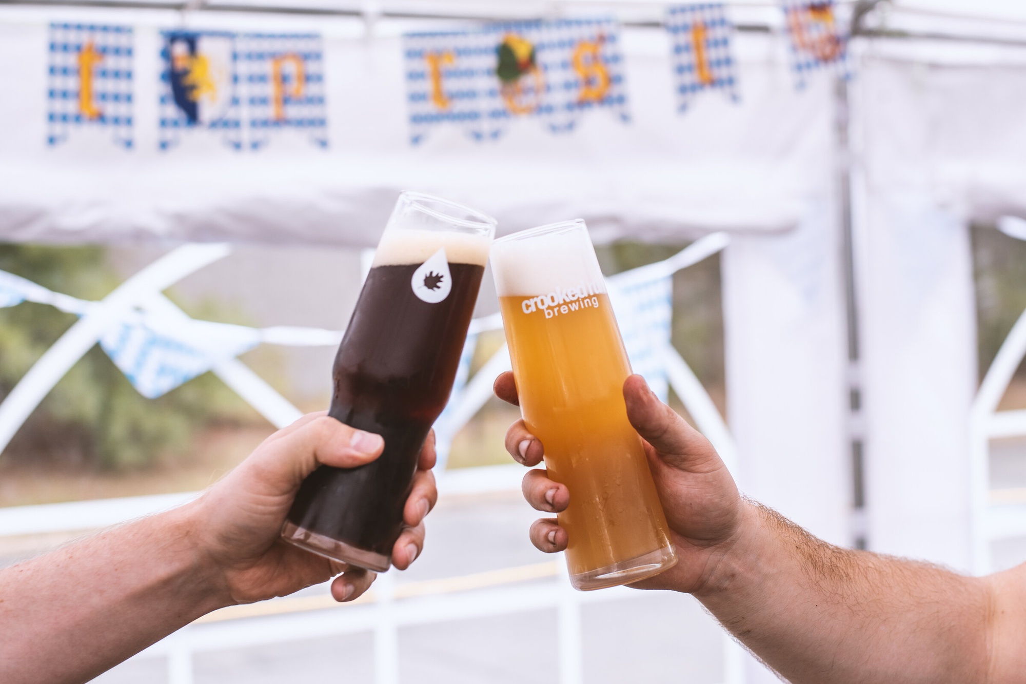 Two hands clink dark and light beer glasses at a festive outdoor setting, with a checkered banner in the background. Casual, celebratory tone.