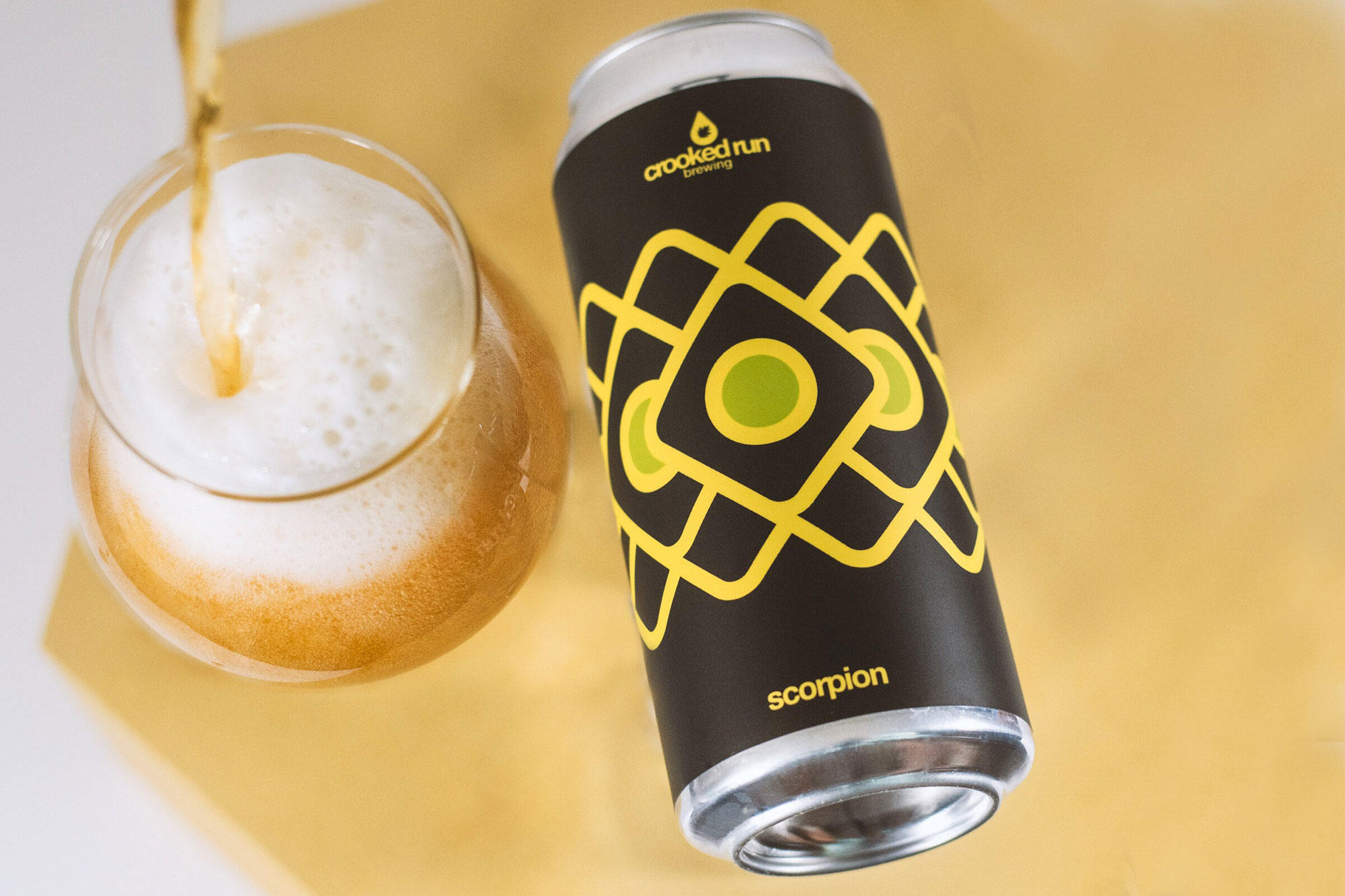 A beer can labeled "Crooked Run Brewing Scorpion" with bold geometric designs is beside a glass being filled with frothy amber beer. The background is golden.