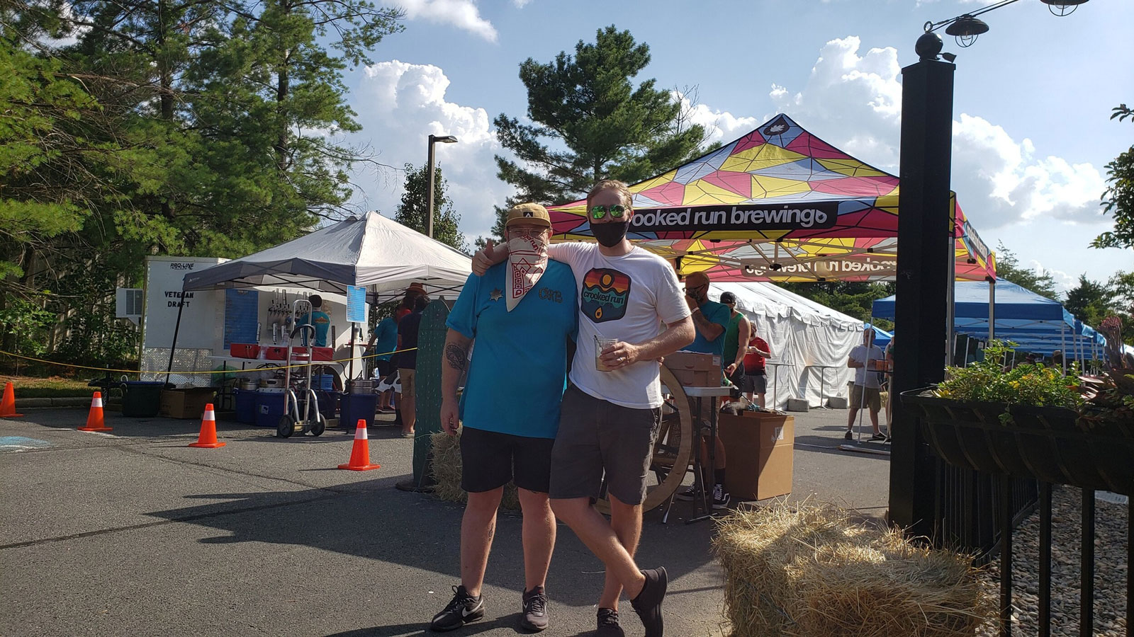 Two people pose happily in front of colorful tents at an outdoor event. Bright sky and trees create a lively, festive atmosphere.