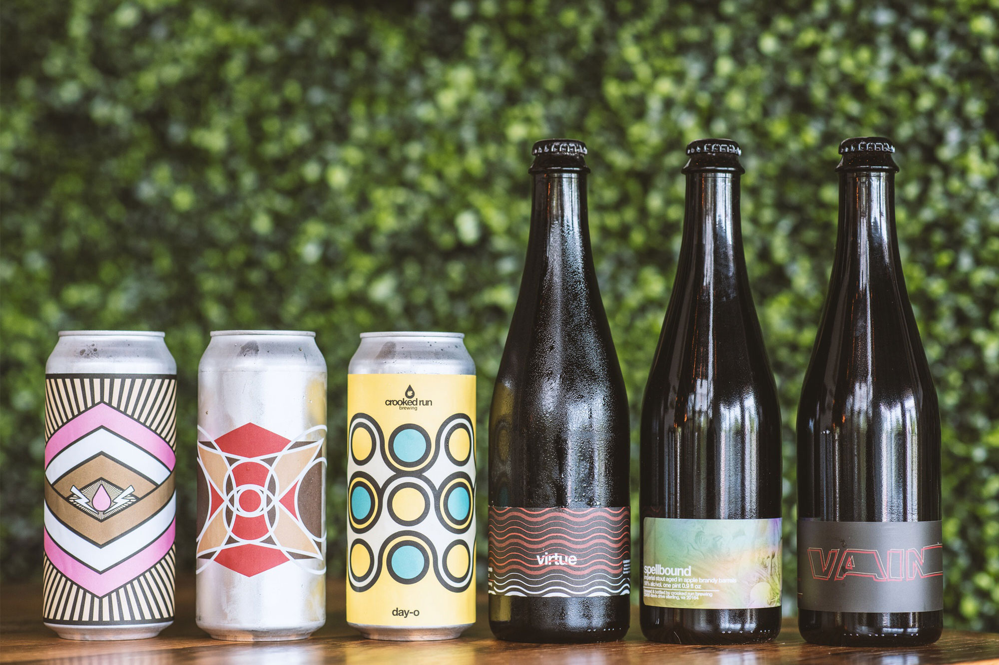 Six craft beer containers, three cans, and three bottles, sit on a wooden surface. Each has a distinct, colorful geometric design, against a leafy backdrop.