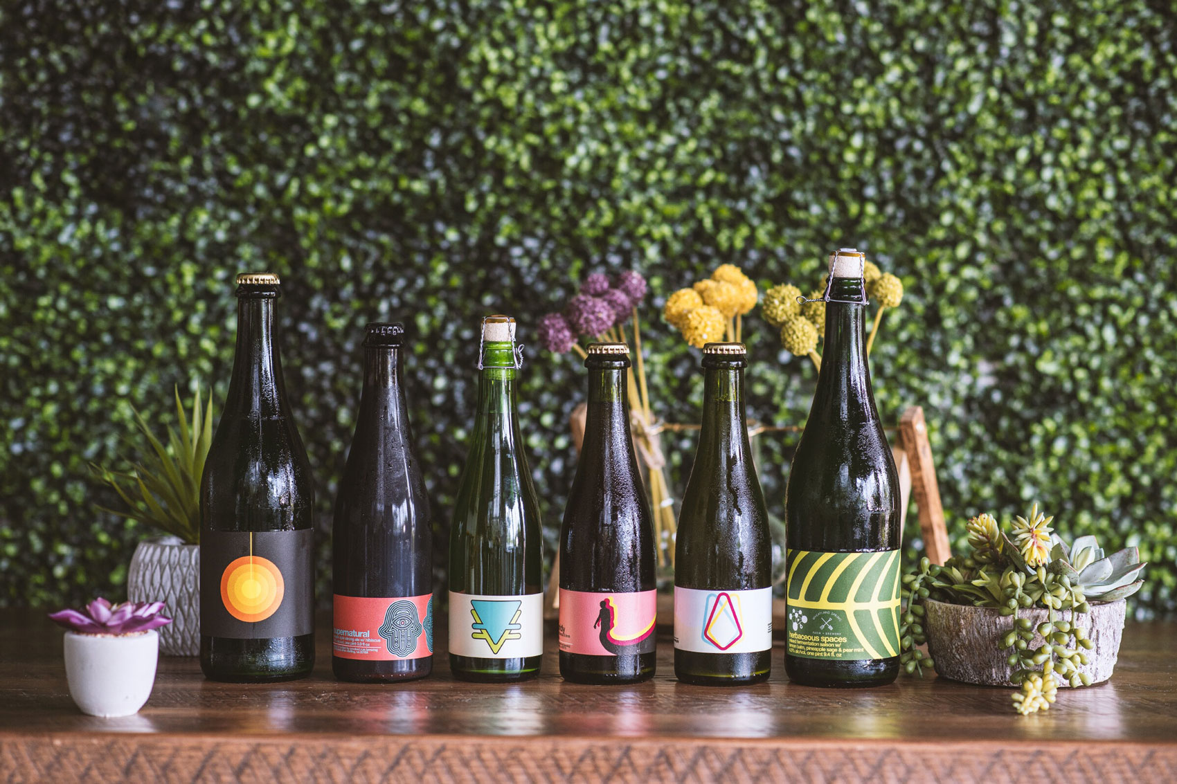 Five uniquely labeled beer bottles on a wooden table with various plants and flowers. A lush green leafy wall serves as the backdrop, creating a fresh ambiance.