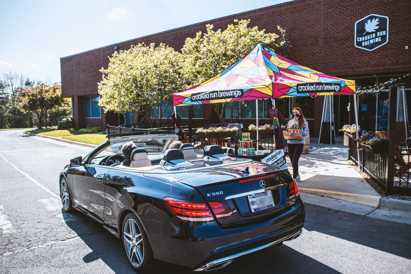 A blue convertible Mercedes approaches a vibrant, colorful tent labeled "Crooked Run Brewing." A woman carries drinks, trees in the background.