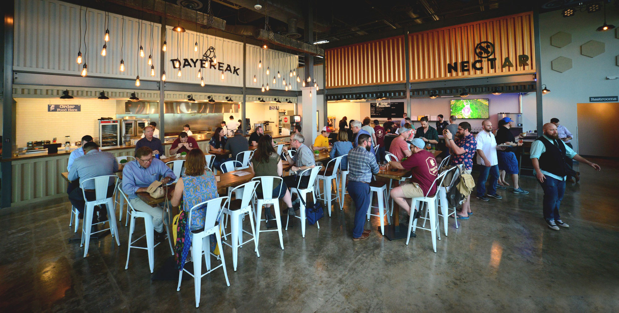 A lively food hall with people socializing at tables. Bright, industrial decor features exposed bulbs and metal signs reading "DAYBREAK" and "NECTAR."