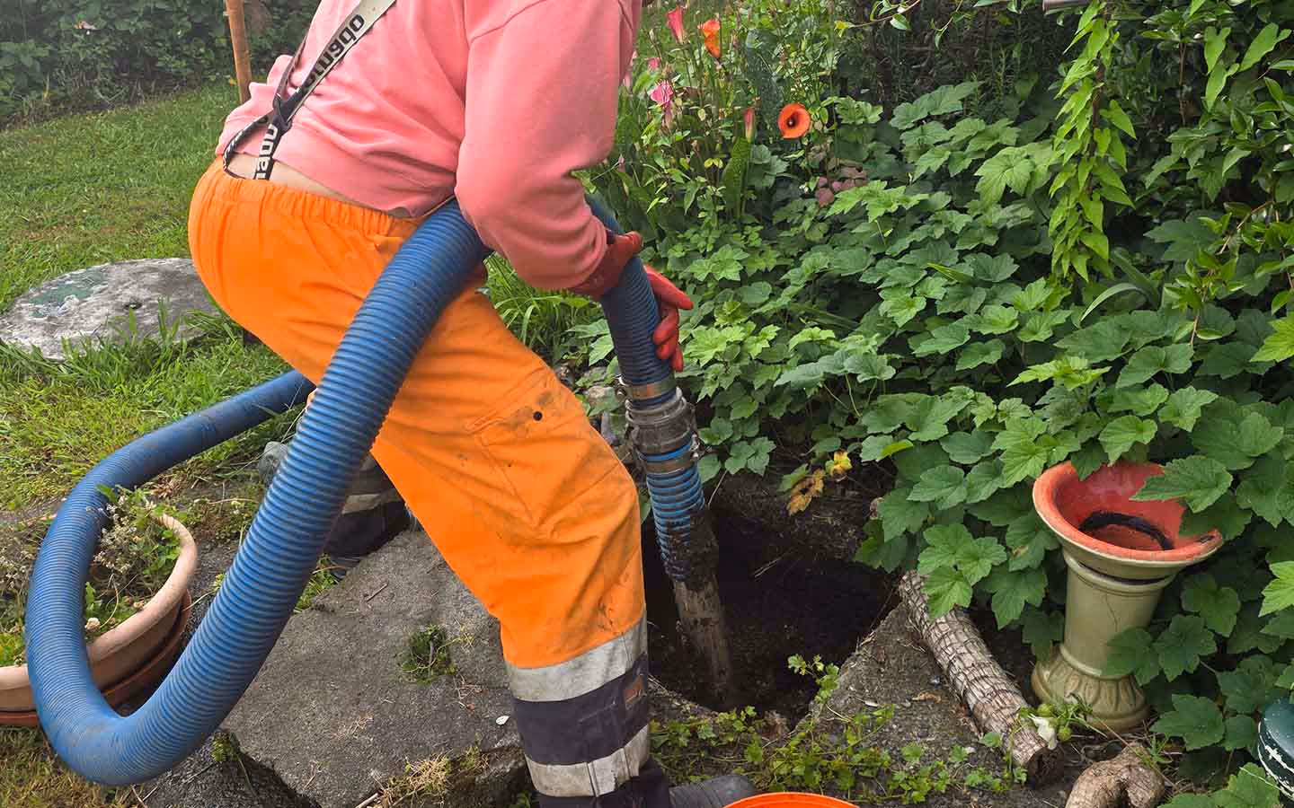 Worker in orange pants and pink top handling a blue hose inserted into a ground hole near green plants and garden pots.