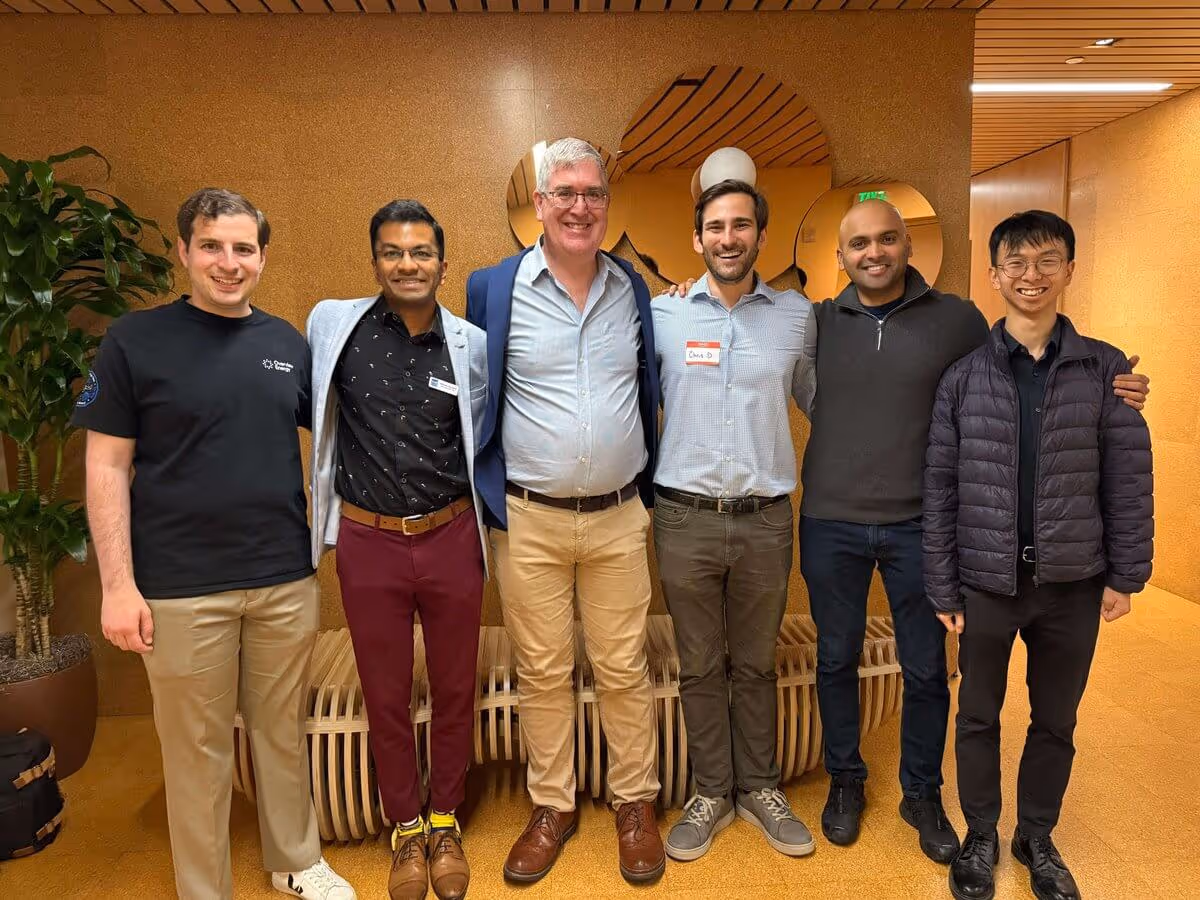 Group of six men standing side by side smiling indoors against a brown wall with a plant nearby.