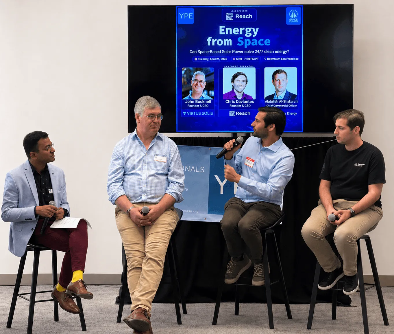 Four men seated on stools in a discussion panel with a screen showing a space solar power event behind them.