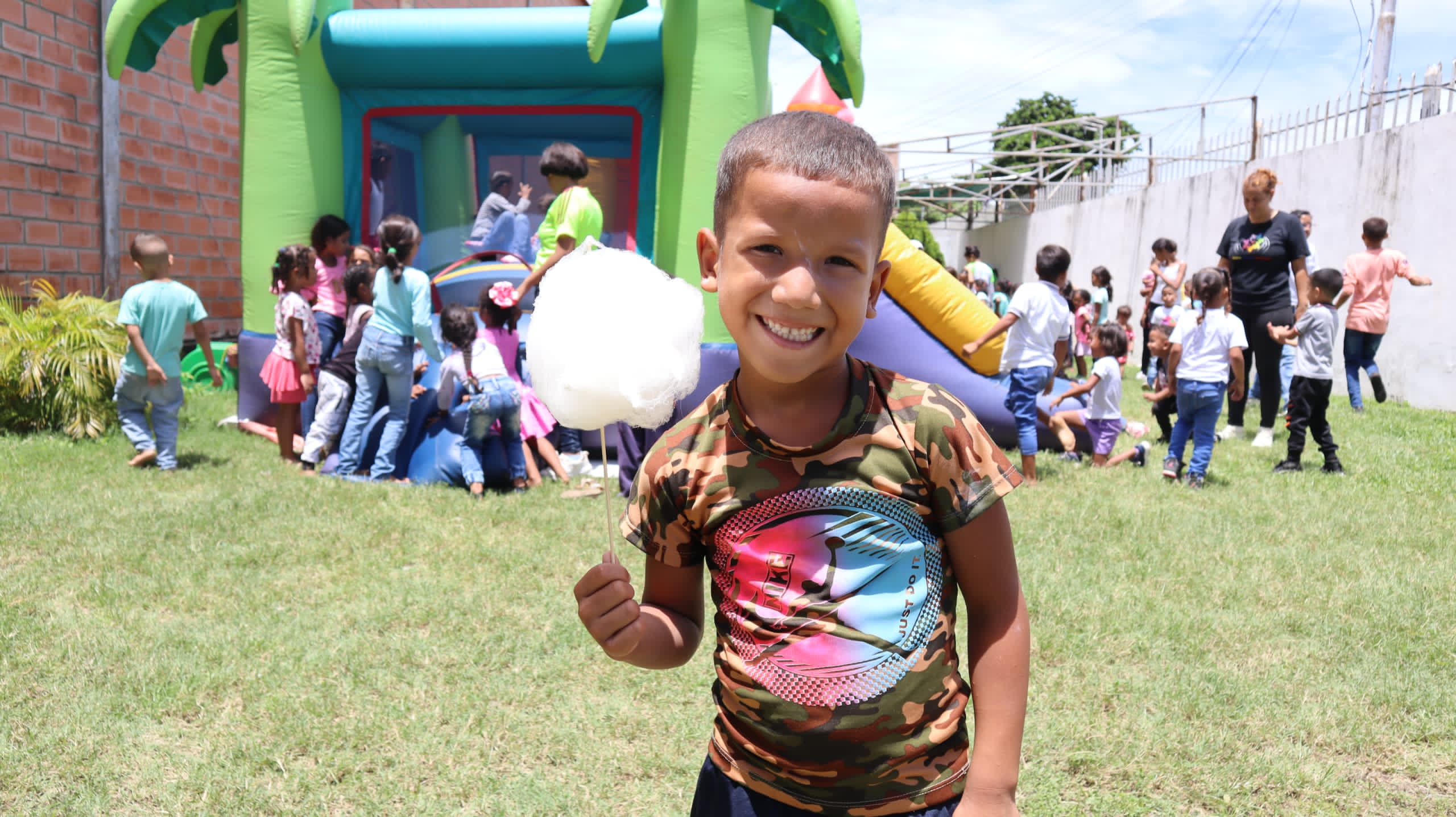 Smiling boy holding cotton candy with children playing near an inflatable bounce house outdoors.