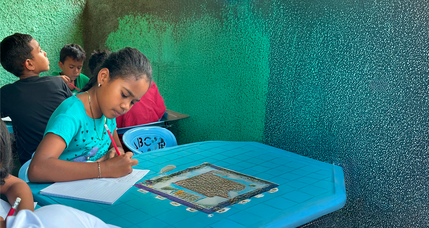 Young girl focused on writing in a notebook at a blue desk in a classroom with other children.