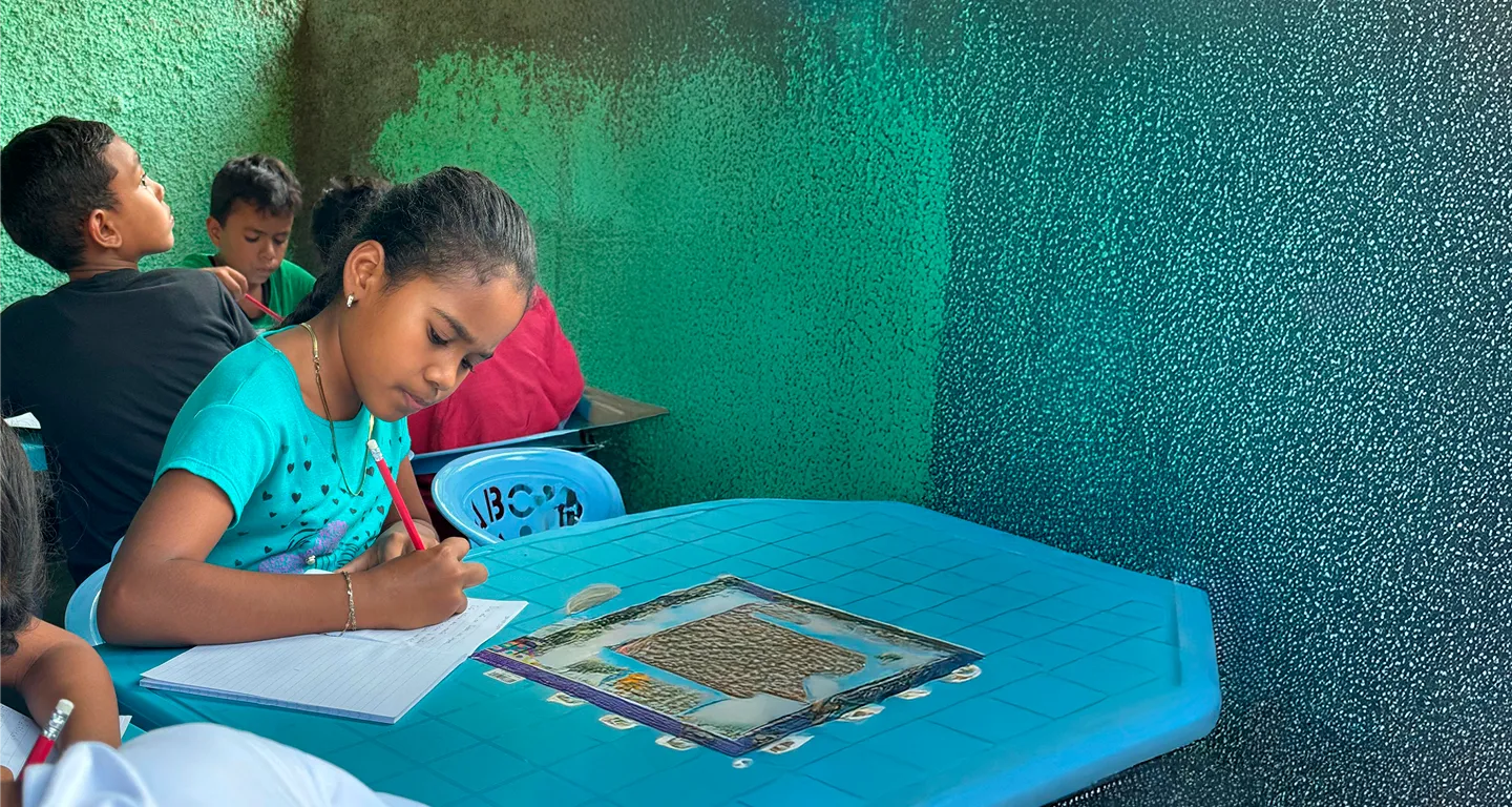 Young girl focused on writing in a notebook at a blue desk in a classroom with other children.