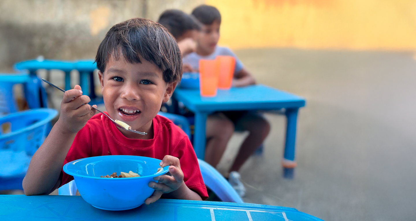 Un niño sonriente come de un tazón azul en una mesa azul, con otros niños detrás.