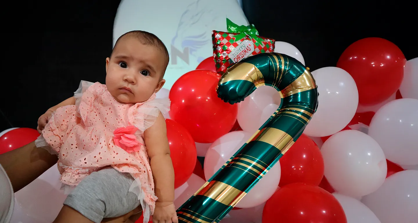 Baby in pink dress held near red and white balloons and a large striped candy cane balloon.