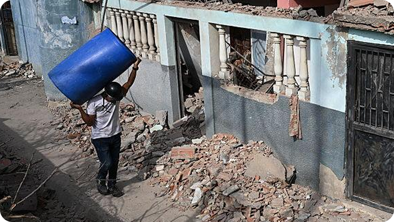 A man carrying a water container past earthquake-damaged houses.