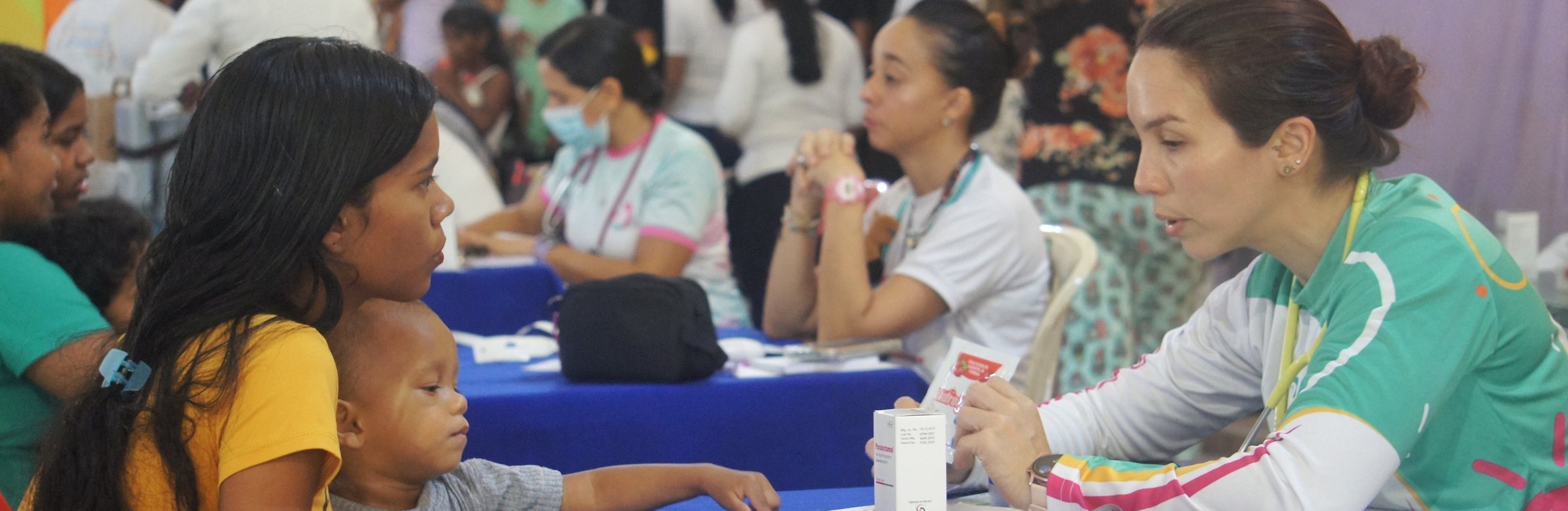 Una madre carga a su hijo en una jornada médica de la Fundación Victoria Venezuela mientras una doctora le explica un medicamento.