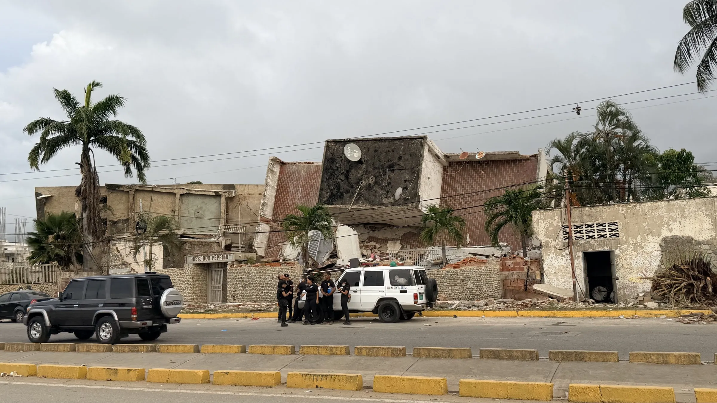 A damaged building on a street, with police officers and a white vehicle in front of it.