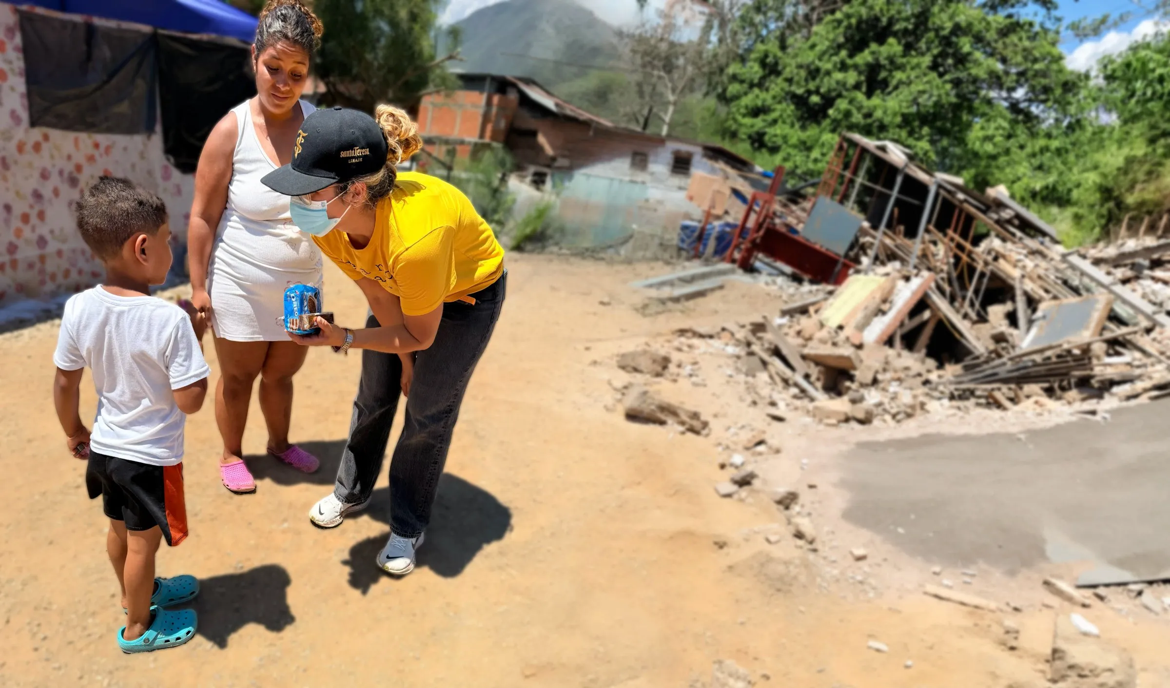 A woman in a yellow shirt hands a drink to a young boy outside a damaged home.
